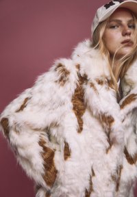 Cappotto in pelliccia sintetica bianco con macchie marroni, caratterizzato da un materiale morbido e peloso e un design ampio. Lo sfondo è di un rosa tenue.