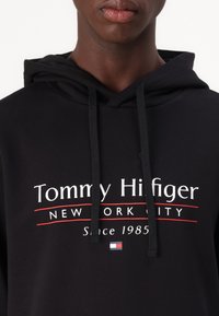 Schwarzer Hoodie aus Baumwolle, mit einem Frontdruck, der "Tommy Hilfiger", "New York City" und "Seit 1985" in weißem und rotem Text zeigt.