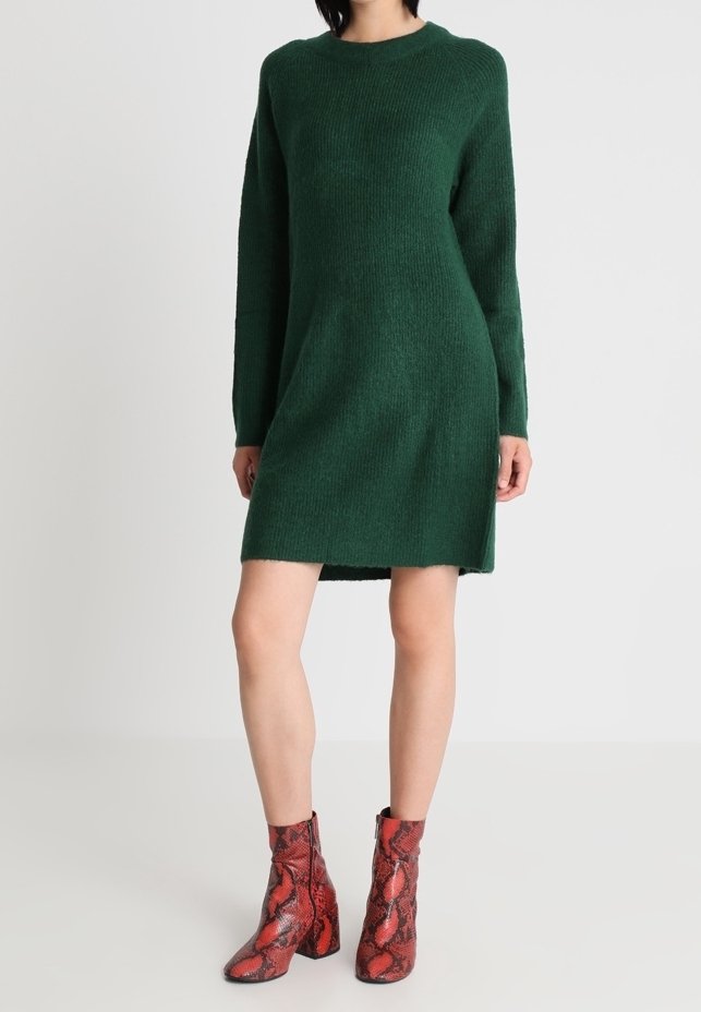 Femme portant une robe en maille verte foncée à manches longues, associée à des bottes mi-mollet à motif peau de serpent rouge, debout devant un fond uni.