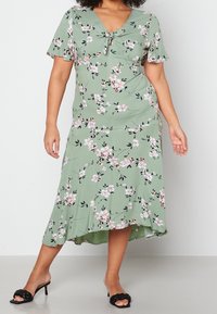 Frau, die ein salbeigrünes Midikleid mit Blumenmuster, kurzen Ärmeln, einem knopfartigen Dekolleté und schwarzen, geflochtenen Absatzsandalen trägt, steht auf einem grauen Boden.