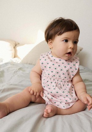 Bébé aux cheveux bruns portant une grenouillère rose à motifs, assis sur un lit avec des draps gris clair et des oreillers beiges.