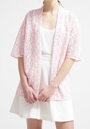 Vrouw draagt een witte gestructureerde rok, wit mouwloos topje en een losse roze bloemen kant cardigan met ellebooglange mouwen.