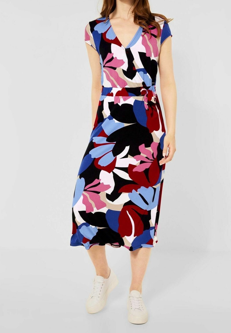 Femme portant une robe midi à motifs floraux colorés avec un décolleté en V et un nœud sur le côté, associée à des baskets blanches décontractées.