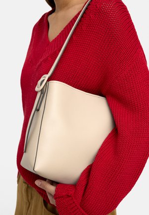 Osoba w czerwonym dzianinowym swetrze trzyma beżową skórzaną torebkę na ramię z okrągłą klamrą na gładkim tle.
