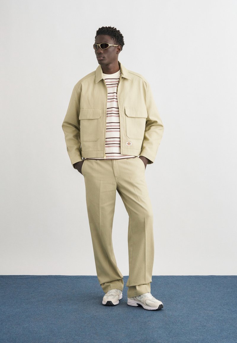 Homme portant une veste et un pantalon beige, une chemise à rayures, des baskets blanches et des lunettes de soleil, debout sur un sol bleu contre un mur blanc.