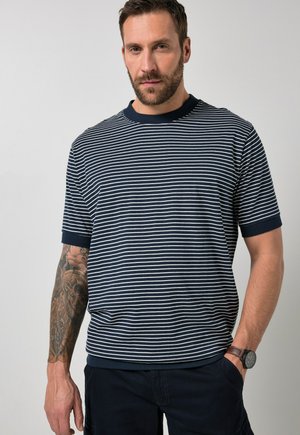 Uomo con capelli castani corti e barba, che indossa una maglietta a maniche corte a righe blu navy e bianca, pantaloni scuri, orologio da polso e tatuaggi visibili sulle braccia.