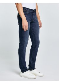 Persoană purtând blugi conici albastru închis și adidași albi, stând pe un fundal gri deschis.