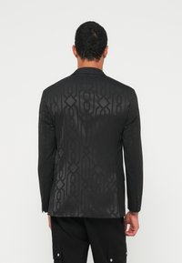 Mees, kes kannab musta bleiserit, millel on peen geomeetriline muster, seisab valge ühevärvilise tausta ees seljaga vaataja poole.