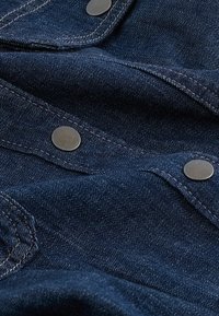 Tumesinine denimkangas nähtava õmblemisega, tekstureeritud pind ja hõbedased metallist nupud. Suurendatud vaade, mis toob esile materjali ja detailid.