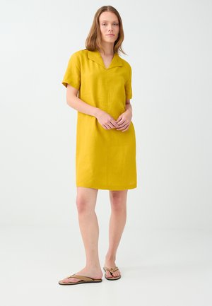 Donna che indossa un vestito giallo senape a maniche corte e sandali beige, in piedi davanti a uno sfondo chiaro e semplice.