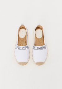 Valkoinen kangaskenkä espadrilles, jossa on kudottu juuttipohja, etuosassa musta nauha, jossa lukee "VALENTINO", ja pyöristetty kärki.