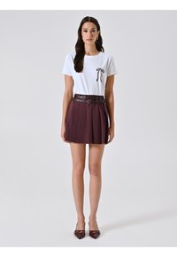 T-shirt bianca con dettaglio a fiocco nero, abbinata a una gonna plissettata bordeaux, arricchita da una cintura doppia nera. Il modello indossa décolleté bordeaux a punta.