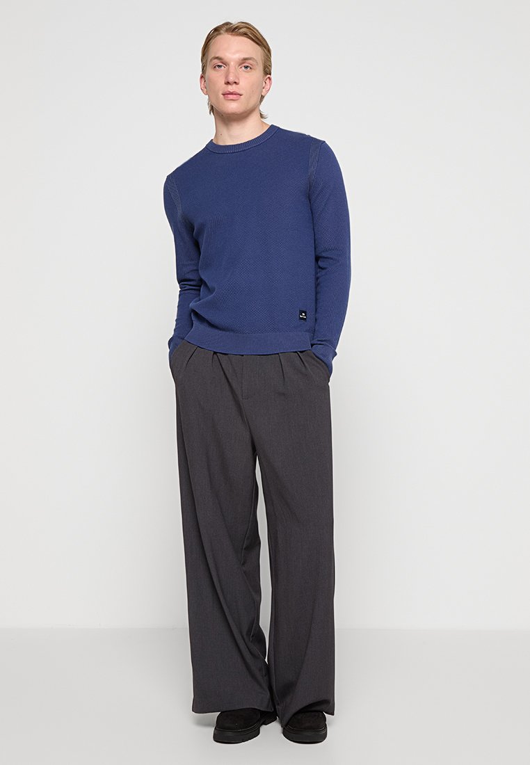 Marinblå stickad tröja med texturerad yta och rund halsringning, kombinerad med grå vida byxor. Svarta accenter finns i klädseln.