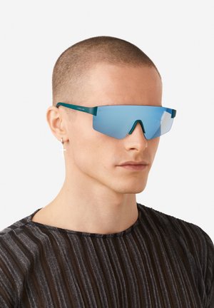 Teal-framed sunglasses feature oversized blue mirrored lenses. The design includes a sharp geometric shape and minimalist metal accents.