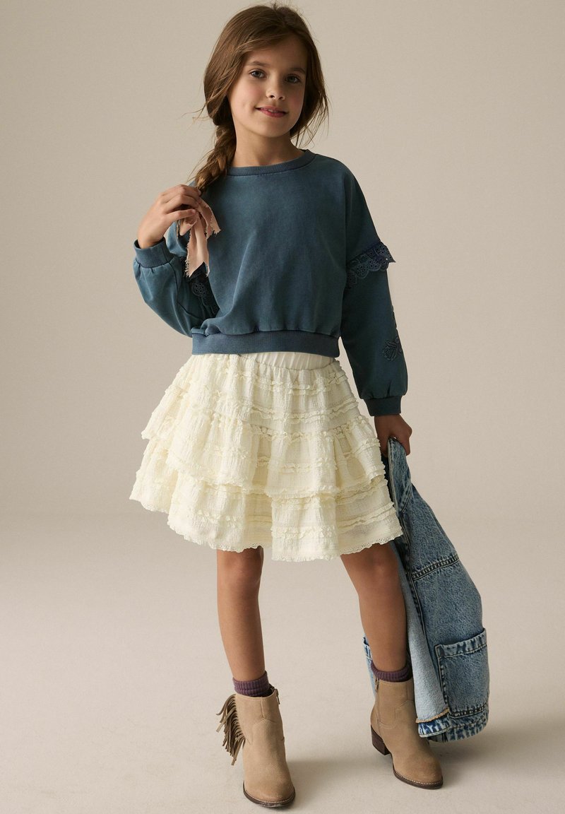 Blauwe sweatshirt met kanten details, gelaagde crèmekleurige rok, tan enkellaarsjes met franjes en een spijkerjack in hand. Zachte texturen, casual stijl.