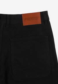 Czarne spodnie z wytrzymałego materiału, z dużą tylną kieszenią i brązową skórzaną naszywką z wytłoczonym napisem "PROSTO".