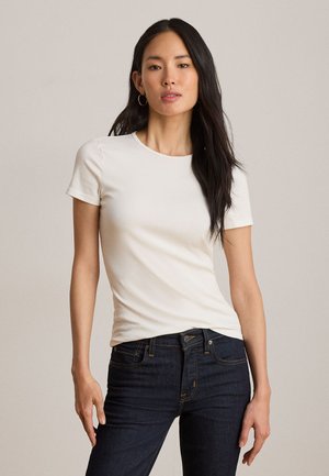 Frau mit langen dunklen Haaren, die goldene Creolen, ein eng anliegendes weißes kurzärmeliges T-Shirt und dunkelblaue Jeans vor einem schlichten Hintergrund trägt.