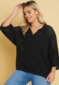 Schwarzer gestrickter Pullover mit lockerer Passform, V-Ausschnitt und ¾ Ärmeln. Verfügt über ein strukturiertes offenes Gewebe und eine Brusttasche. Kombiniert mit blauen Jeans.