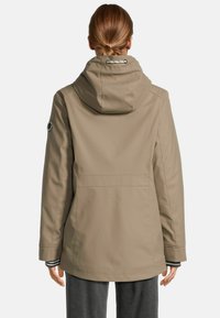 Femme aux cheveux châtain clair relevés en un bas bun, portant une veste à capuche beige et un pantalon gris foncé, se tenant de dos à la caméra.