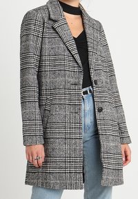 Kvinna i en svart och vit houndstooth-rutigt kappa över en svart topp och ljusblå jeans, med ringar på vänster hand.