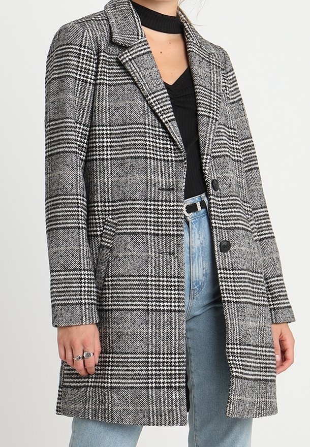 Kvinna i en svart och vit houndstooth-rutigt kappa över en svart topp och ljusblå jeans, med ringar på vänster hand.