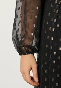 Musta mekko, jossa on läpikuultavat puffihihat, kultaisilla geometrisilla kuvioilla ja joustavilla hihansuilla. Teksturoitu kangas, jossa on sileä aluskangas.