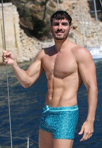 Shorts de bain bleus avec un motif abstrait bleu clair, fabriqués en matériau léger. Un homme se tient près de l'eau, tenant la rambarde d'un bateau, dans un environnement ensoleillé.