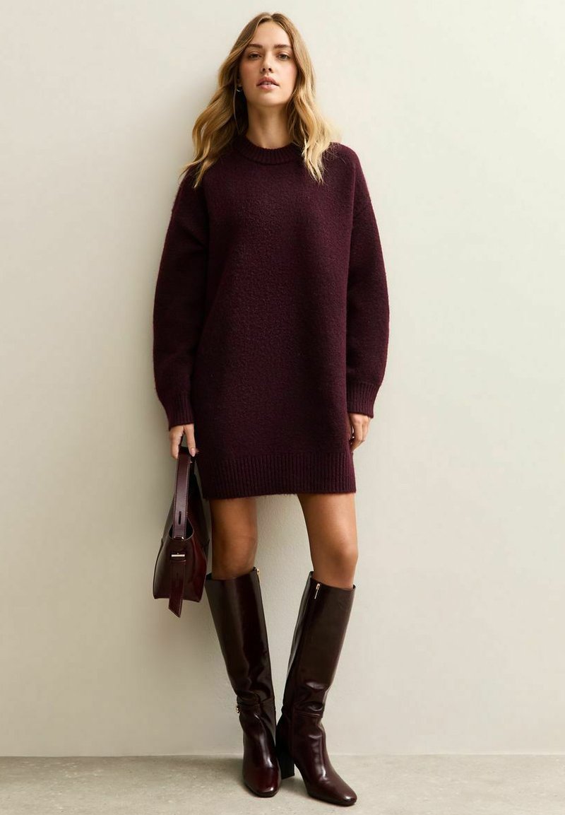 Abito in maglia bordeaux con finitura testurizzata, maniche lunghe e orlo a coste, abbinato a stivali al ginocchio bordeaux lucidi e una borsa coordinata.