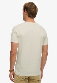 Camiseta de manga corta y cuello redondo en algodón beige claro. Presenta una textura suave y un diseño liso con un pequeño detalle de etiqueta en la parte trasera.