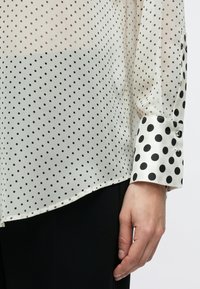 Bluza albă transparentă cu buline negre, prevăzută cu un manșon negru solid cu buline și detaliu de nasture. Textură netedă și croială lejeră.