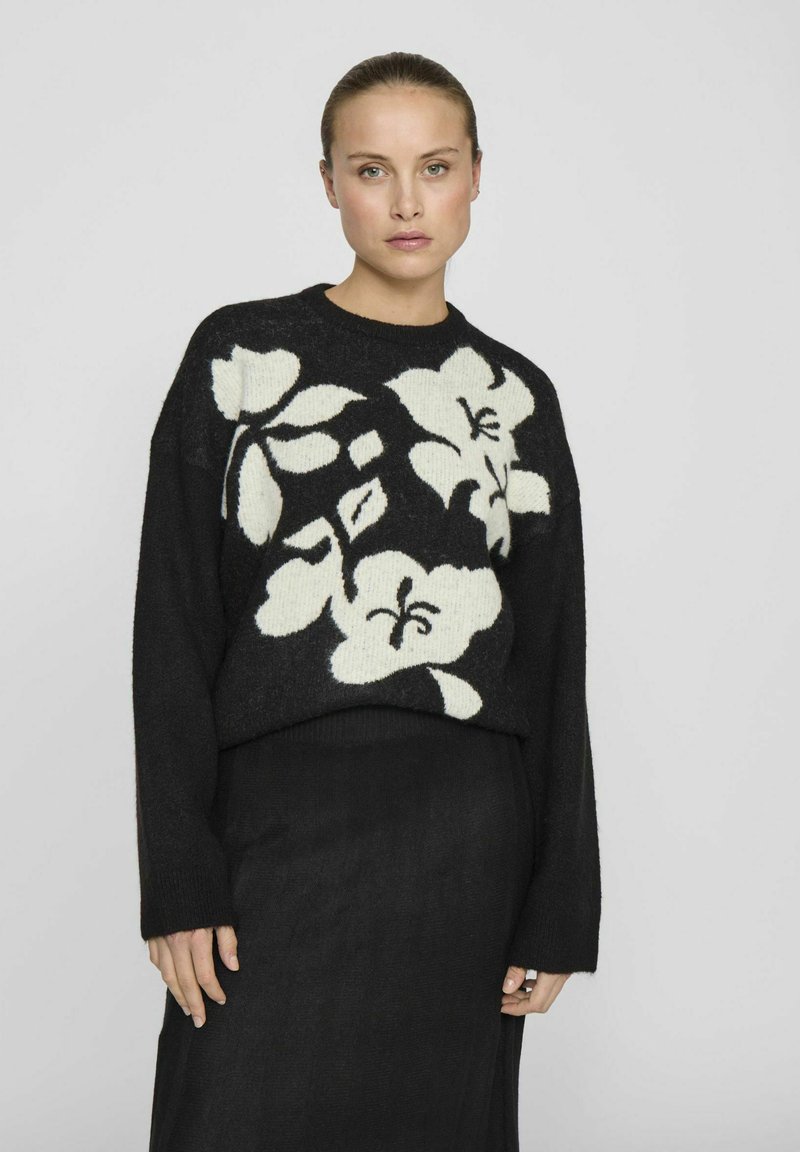 Černý pletený svetr s velkým bílým květinovým vzorem, kulatým výstřihem a dlouhými rukávy. Nosený přes černou sukni z texturované látky.