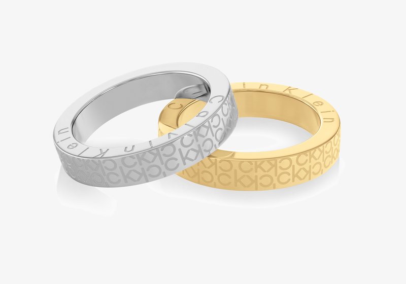 Duas anéis empilhados: um prateado com padrão em relevo "CK", o outro dourado com "Calvin Klein" gravado; formas lisas e redondas com acabamentos polidos.