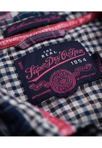 Marinrutigt tyg med guldaccenter; innehåller en framträdande röd och rosa etikett som detaljerar "Superdry" och "British Makers."