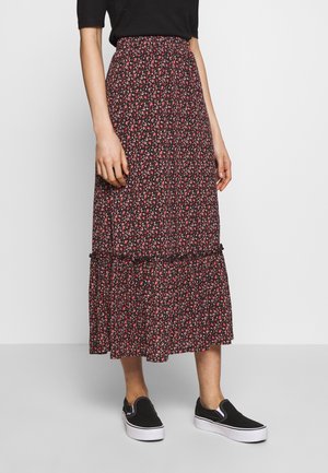 Femme portant une jupe midi fleurie noire avec des fleurs rouges et roses, associée à un haut noir et des baskets slip-on noires à semelles blanches.