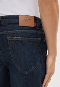 Granatowe dżinsy z ciemnego denimu mają skórzaną łatę z napisem "JOOPI JEANS" na pasku, z pomarańczowym przeszyciem i projektem tylnej kieszeni.
