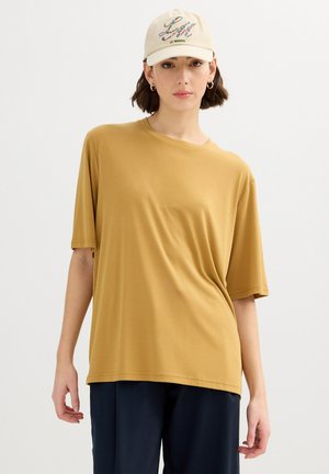 Mujer con una camiseta de manga corta oversize de color mostaza, pantalones azul marino, gorra beige con texto colorido y pendientes de aro dorados, de pie frente a un fondo blanco.