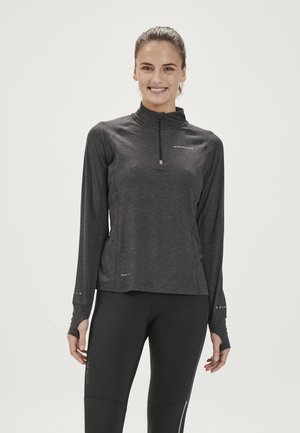 Kvinde smilende, iført en mørkegrå langærmet sportsbluse med tommelhuller og sorte leggings mod en ensfarvet hvid baggrund.