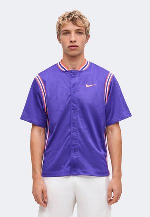 Jeune homme portant un maillot Nike violet à manches courtes avec des détails rayés roses et blancs, debout devant un fond clair uni.