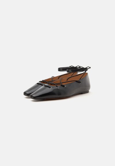 Paire de ballerines en cuir noir brillant avec bouts carrés et fines brides entourant la cheville sur fond blanc.