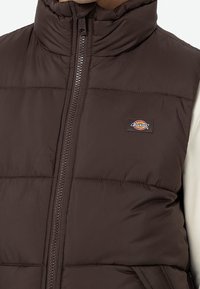 Brun pufferväst med hög krage, dragkedjeslutning och stickade sektioner; har ett litet Dickies-logopatch på bröstet.