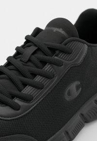 Black sneakers with textured mesh and smooth accents, featuring flat laces, a logo on the side, and a cushioned sole with ridges.