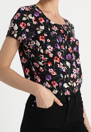 Frau trägt ein schwarzes Kurzarmoberteil mit buntem Blumenmuster und schwarze Hose, Hand in der Tasche, vor schlichtem Hintergrund.