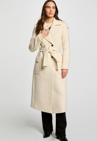 Crème trenchcoat met een ceintuur in de taille, dubbele knopen, zijzakken en een gladde textuur, gecombineerd met donkere broek en laarzen.