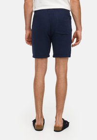 Marineblauwe katoenen shorts met een gestructureerd oppervlak, een elastische tailleband en een achterzak. Eindigt boven de knie. G gedragen met zwarte instappers.