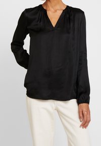 Blusa nera a maniche lunghe realizzata in tessuto morbido. Presenta un collo a V e dettagli arricciati sulle spalle. Indossata con pantaloni chiari.