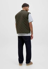 Quilted olivengrøn vest over en grå skjorte, parret med mørke denimjeans og brune sko. Glat tekstur med en afslappet pasform.