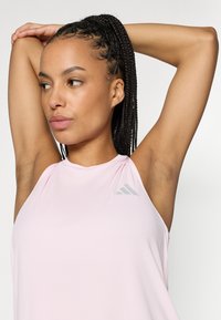 Ροζ αθλητική φανέλα με ανοιχτό ροζ χρώμα, υψηλή λαιμόκοψη, χωρίς μανίκια και γκρι λεπτομέρεια λογότυπου· ύφασμα λείο με ελαφριά γυαλάδα.