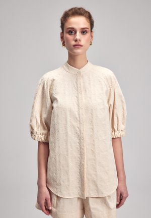 Cremfarbene Bluse mit strukturierten Blumenmustern, Stehkragen und kurzen Puffärmeln. Knopfverschluss vorne; lockere Passform.
