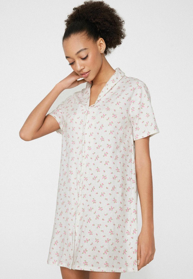 Kobieta o kręconych włosach w białej sukience z krótkim rękawem pokrytej małymi, różowymi kwiatowymi wzorami, stojąca z jedną ręką dotykającą szyi.