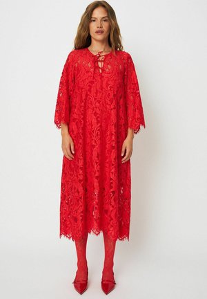 Ženska, oblečena v rdečo čipkasto obleko do kolen s tremi četrtinami rokavi, ujemajoče rdeče vzorčaste nogavice in rdeče čevlje z zoženim prstom.